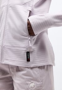 Άτομο που φοράει ανοιχτό γκρι μπουφάν με φερμουάρ και το χέρι στην τσέπη του πλαισίου, καθώς και παντελόνι που ταιριάζει με λογότυπο μαμούθ κοντά στην τσέπη.