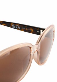 Lunettes de soleil rose clair avec une finition brillante, des montures surdimensionnées et des verres teintés marron. Les branches présentent un motif écaille de tortue avec un marquage "CE".