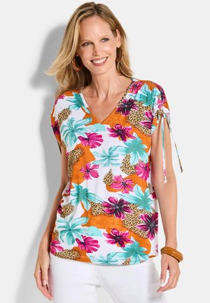 Vrouw lacht en draagt een kleurrijke top met bloemen- en luipaardprint met strikmouwen, witte broek, grote oorringen en een bruine armband.