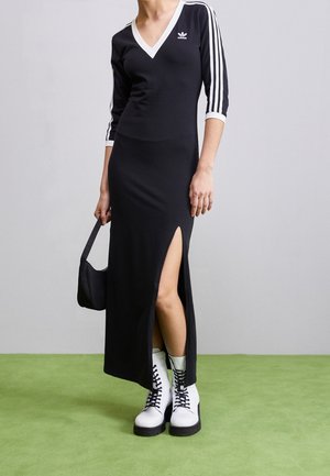 Osoba ubrana w czarną sukienkę Adidas z białymi paskami na rękawach i bocznym rozcięciem, trzyma czarną torebkę i nosi białe sznurowane botki.