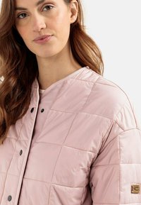 Lys pink quiltet pufferjakke med trykknaplukning, ribbet halsudskæring og et lille logo-patch på venstre ærme. Blød tekstur.