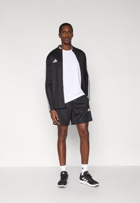 Chaqueta deportiva negra y pantalones cortos, camiseta blanca, calcetines blancos con rayas negras, zapatillas negras. La chaqueta tiene rayas blancas a lo largo de las mangas y un logo.