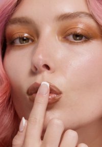 Close-up van een gezicht met glanzende bruine lippen, kort wit-frans gemanicuurde nagels, zachtroze haar en warme oranje oogschaduw.