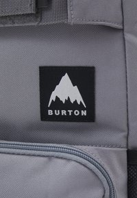 Gray fabric with visible texture features a black patch displaying a white mountain logo and "BURTON" text below. Zipper detail is visible.