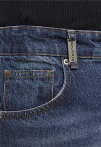 Gros plan sur la poche d'un jean en denim bleu avec couture orange et étiquette métallique portant l'inscription « 2Y STUDIOS » sur la boucle de ceinture, sur fond de tissu noir.