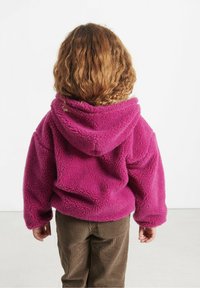 Barn med krøllet hår iført en lys pink fleece hættetrøje og brune fløjl bukser, stående med ryggen til mod en hvid baggrund.