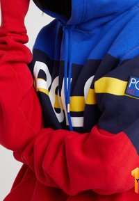 Sudadera roja y azul con un gran logo blanco de "Polo", rayas amarillas y marinas, capucha con cordón y puños acanalados. Tejido suave.