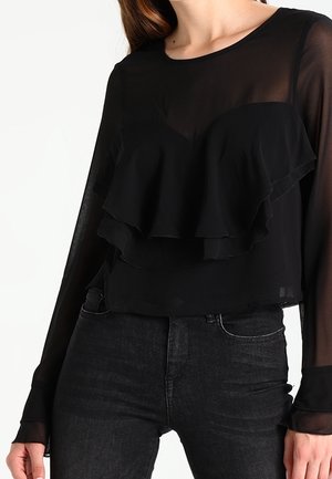 Blouse noire à manches longues avec des volants superposés à l'avant, en tissu transparent. Associée à un jean foncé, créant un look contrasté.