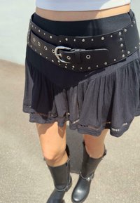 Fustă plisată neagră cu un tiv texturat, echipată cu o curea lata din piele neagră cu ținte metalice și o cataramă argintie, și cizme negre.