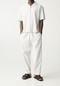 Chemise blanche à manches courtes rayée avec boutons, assortie à un pantalon rayé et des sandales noires, fabriquée dans un tissu doux et léger.