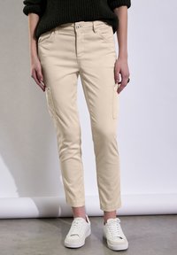 Pantalons cargo beiges en mélange de coton avec poches latérales, coupe slim et ourlets retroussés, associés à des baskets blanches sur une surface gris clair.