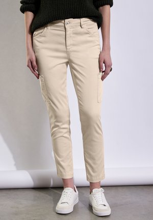 Pantalones cargo beige en mezcla de algodón con bolsillos laterales, corte ajustado y puños, combinados con zapatillas blancas sobre una superficie gris claro.