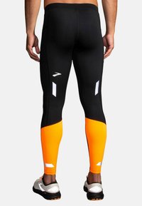 Svarta och oranga träningsleggings med reflekterande detaljer och en åtsittande design, med en gradient från svart till stark orange vid underbenen.