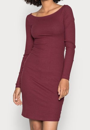 Kobieta ubrana w dopasowaną, prążkowaną, bordową sukienkę z długim rękawem i szerokim dekoltem, pozująca z jedną ręką swobodnie opuszczoną wzdłuż ciała.