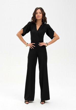 Woman standing with hands on hips wearing a black wide-leg jumpsuit with short sleeves and gold bracelets on both wrists.