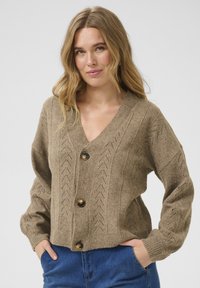 Gilet en tricot beige avec un décolleté en V profond, des poignets côtelés et des motifs en chevron décoratifs. Il est doté de cinq boutons marron sur le devant.