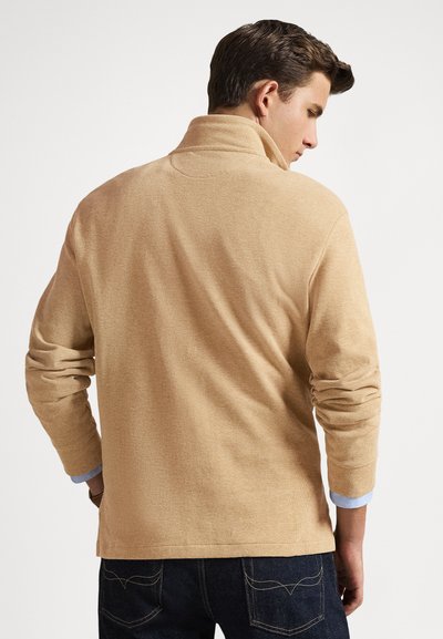 Muž s krátkými hnědými vlasy, oblečený v béžovém svetru a tmavých džínách, otočený zády, s mírně vyhrnutými rukávy, na bílém pozadí.