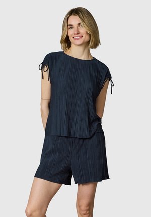 Jeune femme souriante, portant un top court plissé bleu marine avec des détails à nouer et un short plissé assorti.