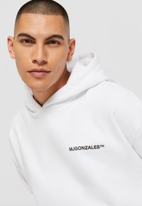 Hvid hættetrøje lavet af blød stof med et frontlogo "MJGONZALES™" i sort. Designet inkluderer en hættetrækning og ribbede manchetter.