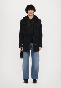 Zwarte jas met meerdere zakken, cinched taille, knoopsluiting. Draagover een zwarte, gestructureerde shirt; gecombineerd met relaxte blauwe jeans en zwarte schoenen.