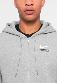 Grijze zip-up hoodie met trekkoorden, zachte textuur en geborduurd Nike-logo. Bevat bedrukte tekst: "ATHLETIC DEPT JUST DO IT." Zwart binnenlaag zichtbaar.