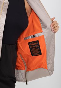 Giacca beige aperta che mostra la fodera arancione brillante con una tasca etichettata "Bombers Original" e una striscia riflettente sopra la tasca con bottoni.