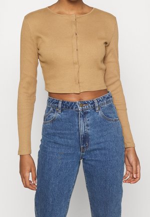 Mujer con un top corto de manga larga acanalado color marrón claro y vaqueros azul denim de talle alto, de pie contra un fondo claro y liso.