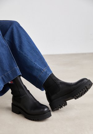 Stivaletti neri in pelle con pannelli laterali elasticizzati, suole spesse a piattaforma e grip testurizzato. Abbinati a pantaloni in denim blu larghi.