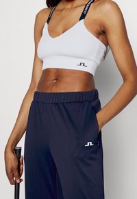 Bílá sportovní podprsenka s žebrovanou texturou a výstřihem do V, černými ramenními popruhy. Námořnicky modré kalhoty s elastickým pasem a logem na bocích.