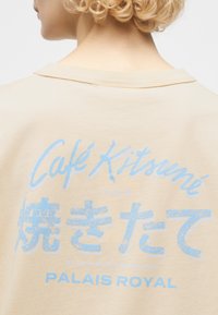 Άτομο με ξανθά, σγουρά μαλλιά, που φοράει μπεζ πουκάμισο με μπλε κείμενο που γράφει «Café Kitsuné», ιαπωνικούς χαρακτήρες και «Palais Royal» στην πλάτη.