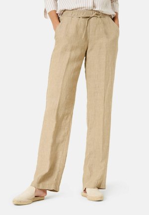 Persona con pantalones holgados de lino beige con cintura ajustable y zapatos sin cordones color crudo, de pie con las manos en los bolsillos.