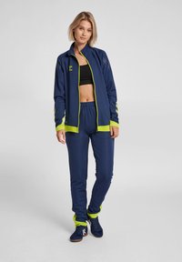Chándal deportivo color marino con acentos en amarillo neón. Cuenta con una chaqueta con cremallera, mangas largas y pantalones con cinturilla elástica. Incluye una camiseta corta negra.