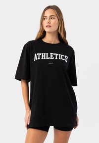 Liela izmēra melns T-krekls, izgatavots no kokvilnas, ar treknām baltām uzrakstu "ATHLETICS" priekšpusē un īsām piedurknēm.