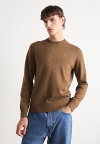 Bruine gebreide trui met een ronde hals en geribde boorden, met een klein vierkant logo op de borst. Draagt met blauwe spijkerbroek.