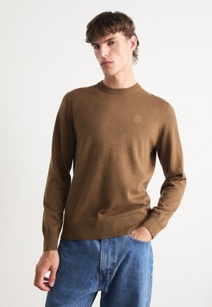 Pullover marrone in maglia con scollo rotondo e polsini a costine, caratterizzato da un piccolo logo quadrato sul petto. Indossato con jeans blu in denim.