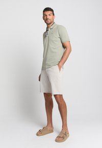 Lichtgroen polo shirt, beige broekjes en tan sandalen. De polo heeft een gestreepte nek sjaal. Glad textuur, casual pasvorm.