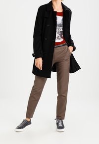 Zwarte trenchcoat met knoopsluiting, rode graphic t-shirt, bruine op maat gemaakte broek en grijze sneakers met witte zolen.