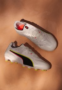 Pari valkoisia Puma-jalkapallokengän nastoja, joissa on mustia ja keltaisia raitakoristeita, vaaleanpunainen vuori ja muotoillut nastat vaaleanruskealla pinnalla.