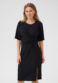 Robe noire mi-longue à manches courtes, avec un détail de devant torsadé et une fente sur le côté. Le tissu texturé offre un motif subtil.