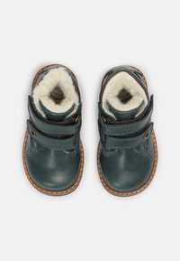 Botas de cuero verde con dos correas de Velcro, forro de pelo y suelas de goma marrón. Presentan una puntera redondeada y detalles de costura.