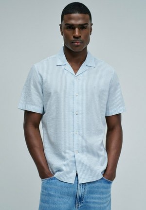 Jeune homme debout, les mains dans les poches, portant une chemise bleu clair à manches courtes boutonnée et un jean en denim sur un fond uni.