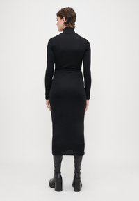 Schwarzes Langarmkleid mit hohem Kragen, figurbetontem Schnitt und knielangem Saum. Das Material wirkt glatt und wird mit kniehohen schwarzen Stiefeln getragen.