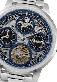 Automatisch horloge met een zilveren roestvrijstalen behuizing, blauwe accenten en blootliggende mechanismen. Beschikt over dubbele tijdsdials en complexe gouden tandwielen.