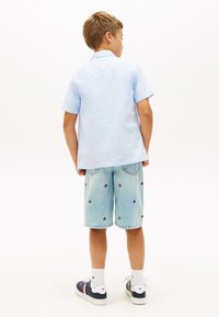 Αγόρι που στέκεται με την πλάτη, φορώντας ανοιχτόμπλε κοντομάνικο πουκάμισο, ανοιχτόχρωμα τζιν σορτς με μικρά λογότυπα, λευκές κάλτσες και ναυτικές αθλητικές παπούτσια.