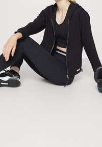 Felpa nera con zip e tasche laterali, abbinata a un reggiseno sportivo nero e leggings. Sneakers nere con suola testurizzata e un kettlebell nelle vicinanze.
