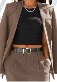 Kobieta ubrana w brązowy garnitur, z marynarką zarzuconą na ramiona, czarny krótki top z prążkowanej tkaniny, czarny pasek z srebrną klamrą oraz warstwowe srebrne naszyjniki.