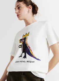 T-shirt en coton blanc arborant une illustration colorée d'un dinosaure portant une couronne, accompagnée du texte "JEAN-MICHEL BASQUIAT."