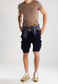 Muž stojící s rukama na bocích, oblečený v hnědém tričku, námořnických cargo šortkách, béžových teniskách a s námořnickým vzorovaným svetrem uvázaným kolem pasu.