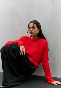 Maglione rosso lavorato a maglia con maniche testurizzate, abbinato a pantaloni neri larghi. Il modello è seduto contro un muro grigio chiaro, mettendo in risalto i colori dell'outfit.