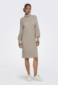 Nais, kes kannab beeži kõrge kaelusega kudumkleiti, millel on pallisülesed varrukad ja valged spordijalatsid, seisab ühetaolise taustaga.